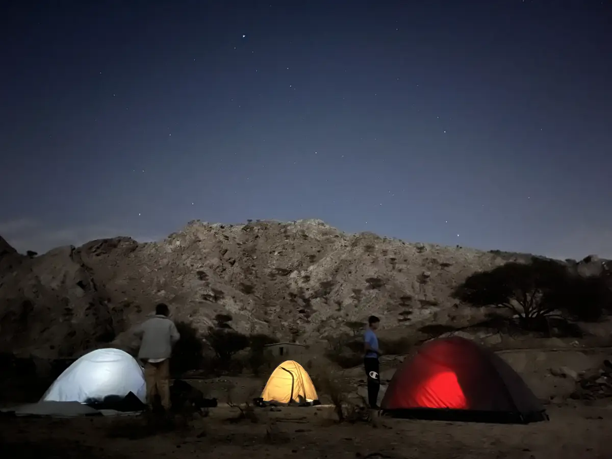 Three tents glow white, yellow and red against dark mountains under a starry night sky