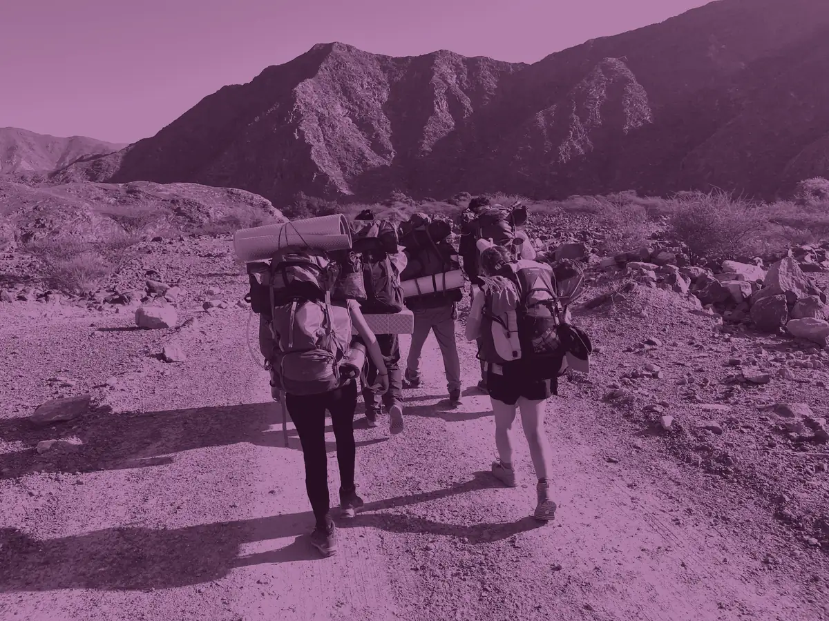 A team with full expedition packs walks away up the trail towards the mountain, washed in Gold purple