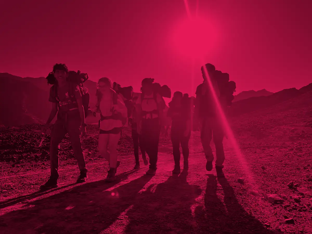 NLCS Dubai students carrying full expedition packs walk out of the sun on a mountain track, washed in the Award's red