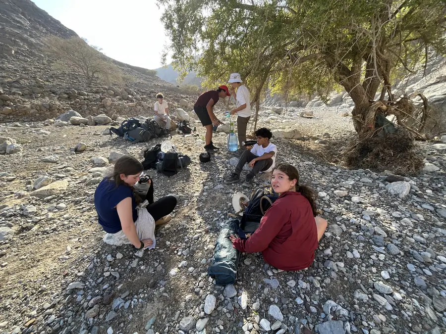 Students rest in the shade of a tree on a stony river bed, packs on the ground