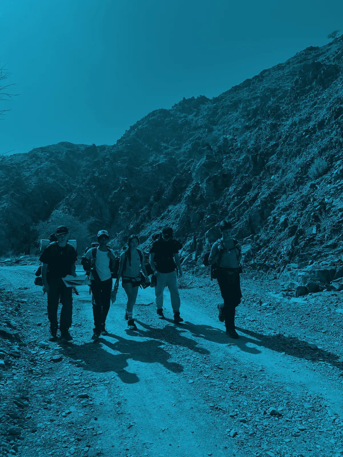 An expedition team walks a stony mountain track beneath a steep ridge line, washed in Silver blue
