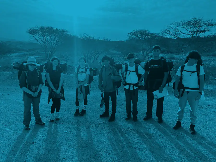 Six students line up shoulder to shoulder in expedition kit, long shadows stretching towards the camera, washed in Silver blue